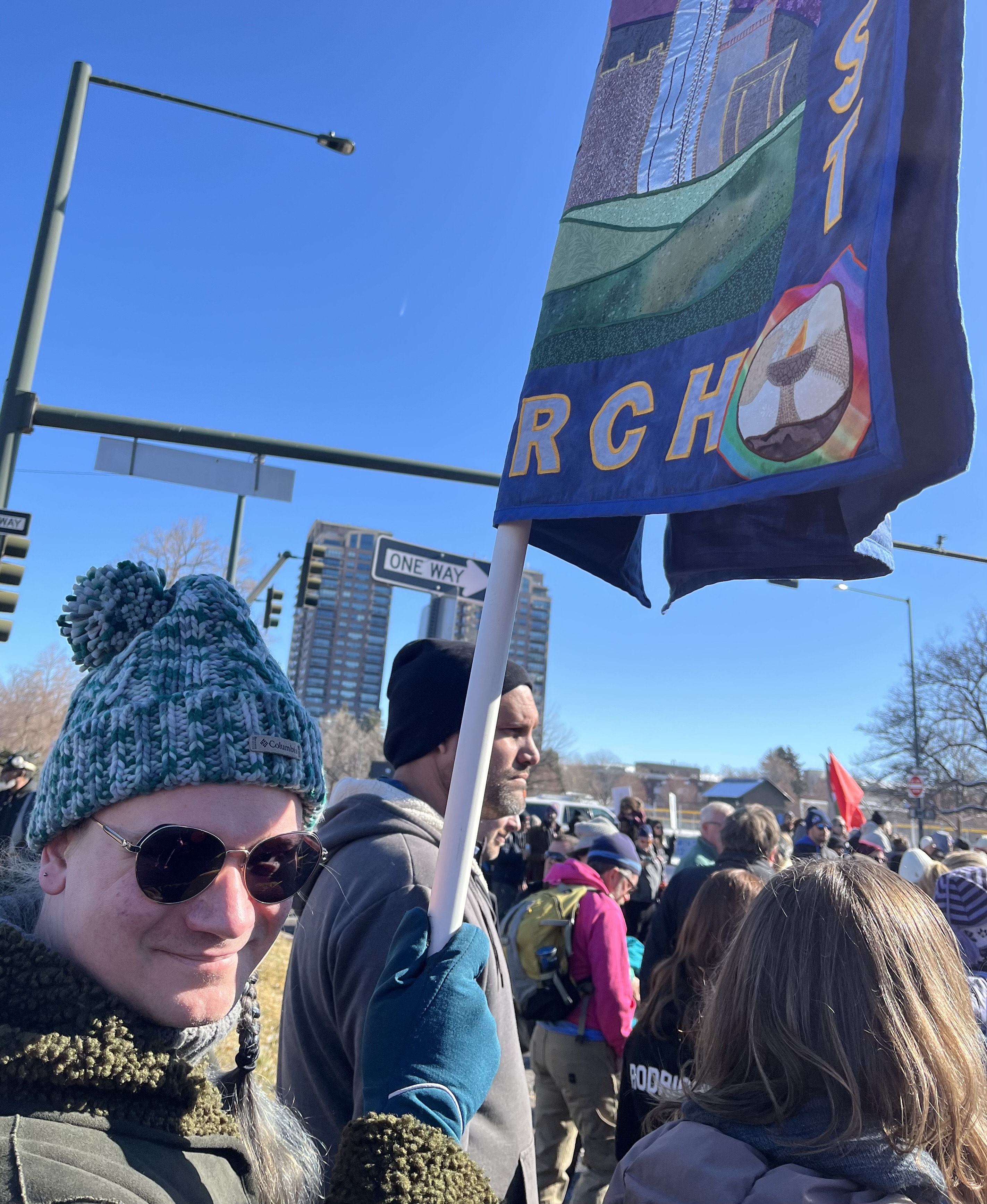 Birch marching with congregants in Denver's annual Martin Luther King Jr. Marade. They are wearing a blue stocking-hat and gloves, a dark green winter coat, and big sunglasses. They are smiling and looking toward the camera. They are holding the church's quilted banner, the bottom of which is visible in the photo.