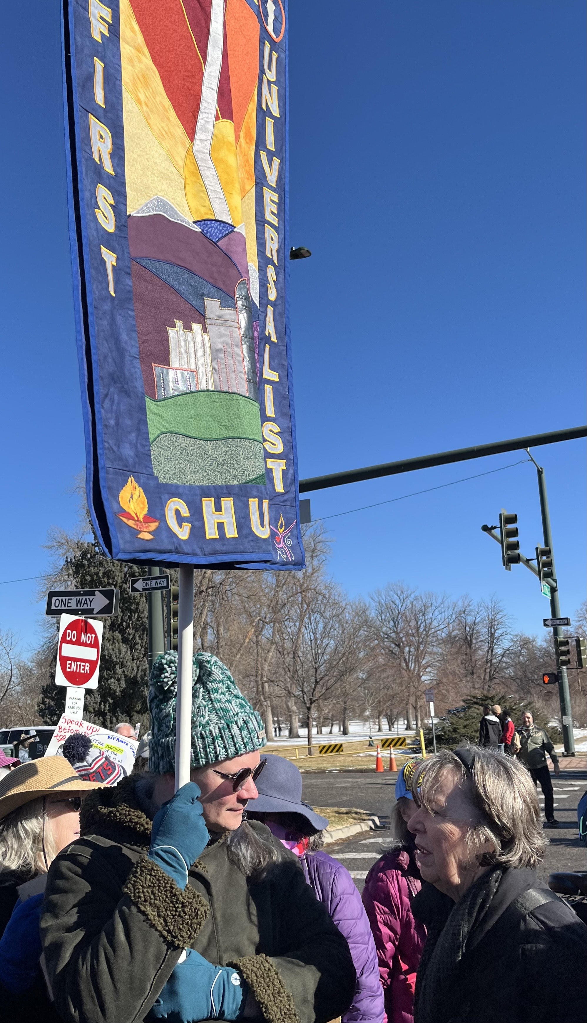 Birch marching with congregants in Denver's annual Martin Luther King Jr. Marade. They are wearing a blue stocking-hat and gloves, a dark green winter coat, and big sunglasses. They are looking at and talking with a congregant, who is visible in profile. They are holding the church's quilted banner, most of which is visible in the photo, including the words FIRST UNIVERSALIST CHURCH. The banner is appliqued with bright fabric, in the shape of the Denver skyline, mountains, and sunset.
